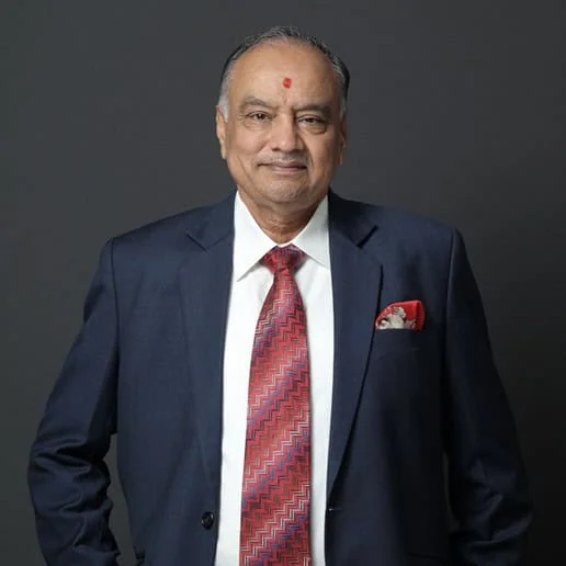 Rajnikant S. Ajmera standing in a navy suit and patterned red tie, photographed against a dark studio background